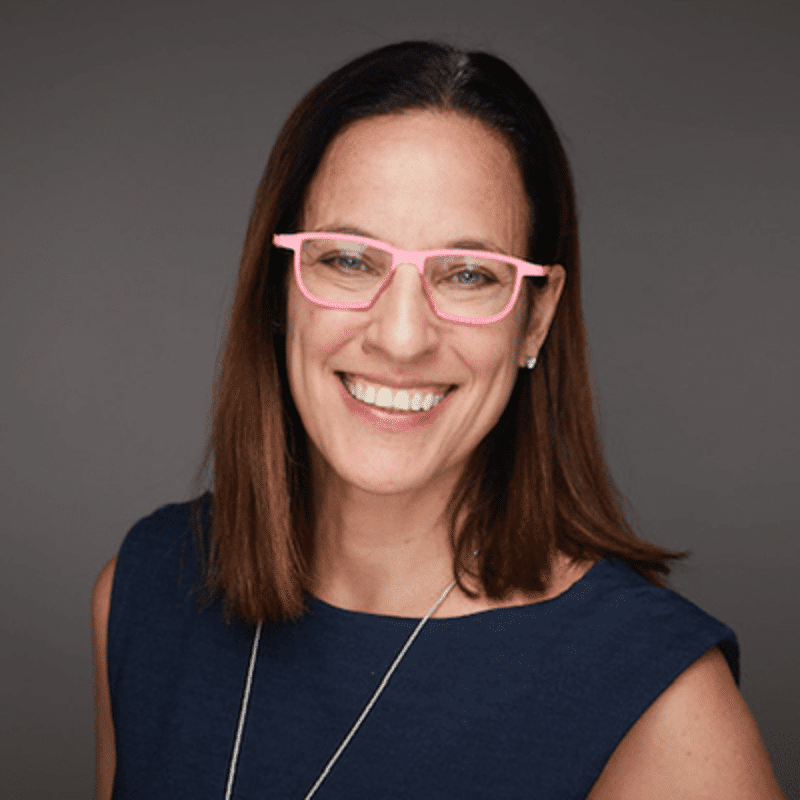 The image shows a woman with brown hair and glasses. She is wearing a dark blue top and a necklace. She is smiling at the camera. The background is a solid gray color.
